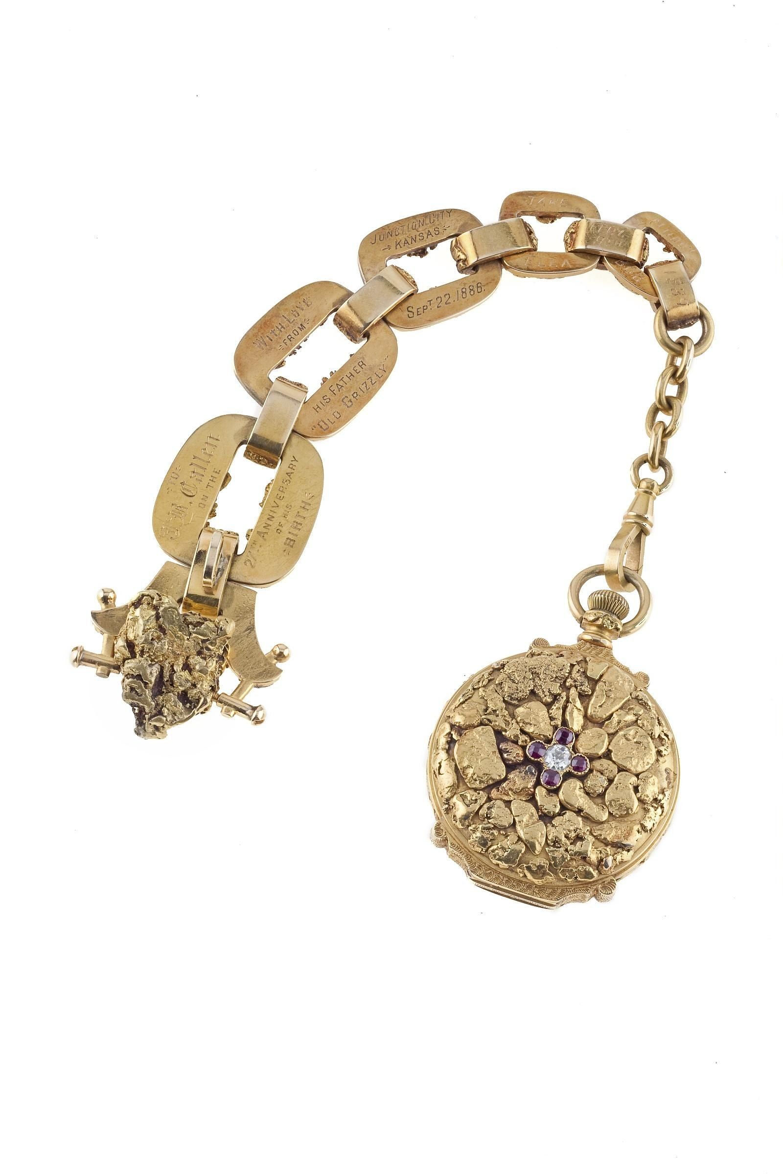 A gold pocket watch covered in natural gold nuggets around a ruby and diamond flower, next to an engraved watch chain ending in a large nugget