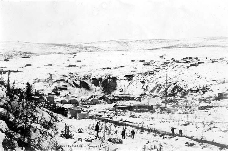Black and white photograph of a snow-covered valley floor dug with mine workings and cabins, a few figures walking along a track