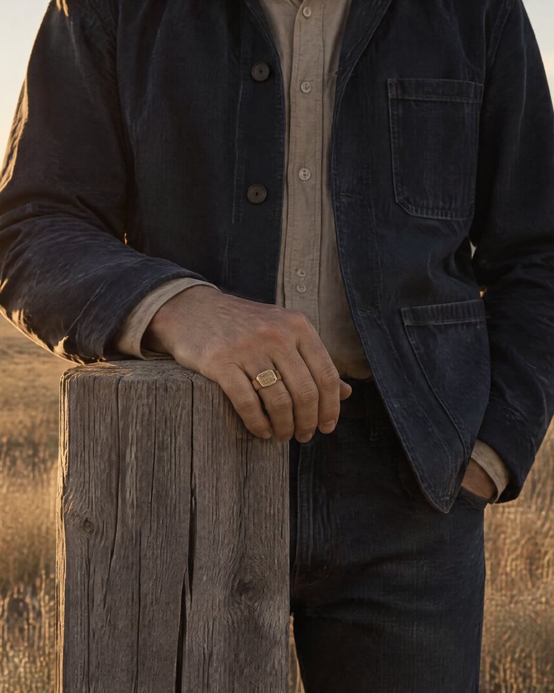 A hand resting on a weathered timber claim post in dry grass at sundown, wearing The Block nugget signet, No. 014, in 14K yellow gold