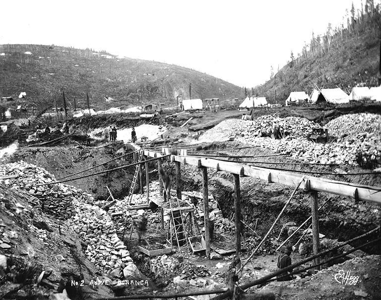 Black and white photograph of a creek valley dug into pits and spoil heaps, with a long wooden flume on trestles and tents behind
