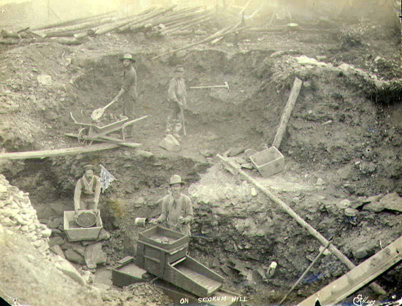 Sepia photograph of four miners in a dug-out pit, two of them working wooden rocker boxes, others with a shovel and a wheelbarrow