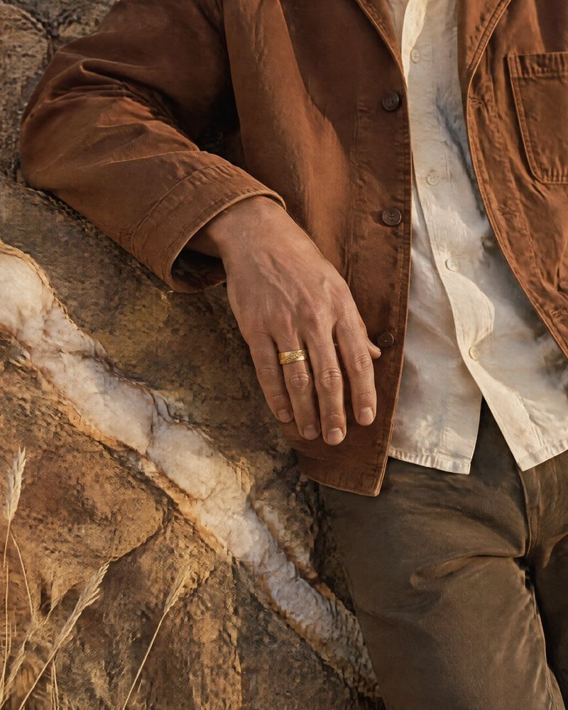 A hand on an ochre rock face crossed by a white quartz vein, wearing the No. 518 Fault Line nugget ring in 14K yellow gold