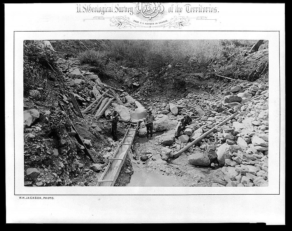 Three men in a rocky gulch working beside a long wooden sluice that runs down through the stones