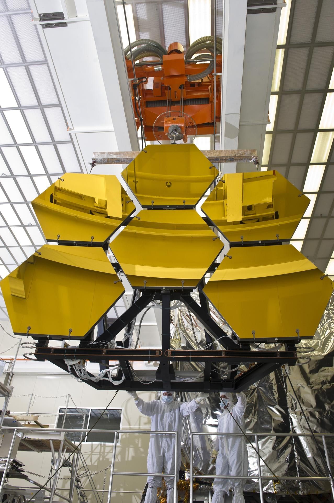 Six gold hexagonal mirror segments mounted together on a black frame, hanging from a crane in a large test hall, with two engineers in clean-room suits guiding it